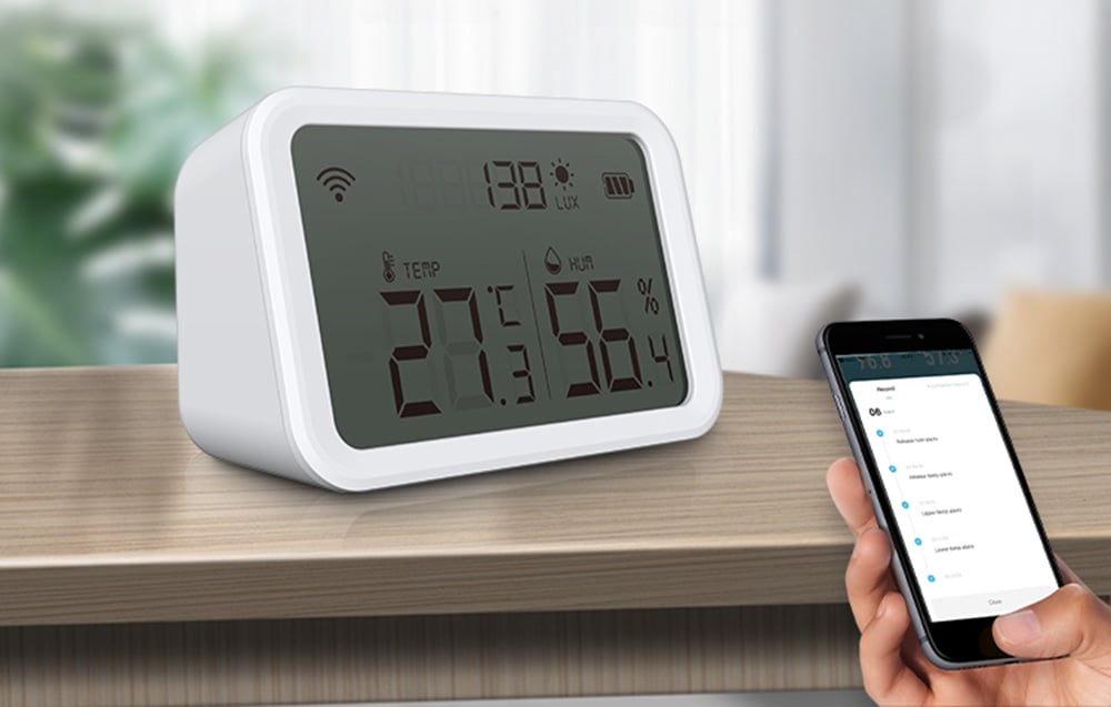 Capteur de température et d'humidité NEO NAS-TH02W avec affichage Zigbee TUYA sur une surface marron. Une main tenant un smartphone est visible.